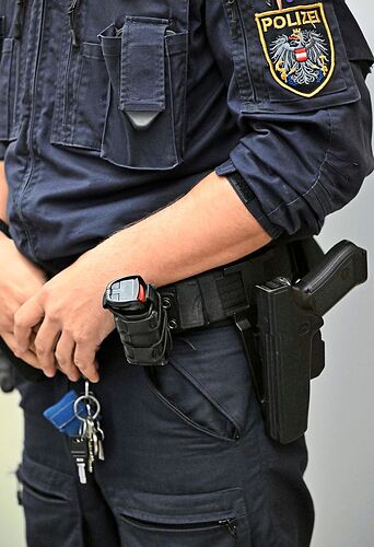 Ein Polizist in Uniform mit Ausrüstung, darunter eine Schusswaffe und ein Taser am Gürtel. Außerdem trägt er Schlüsselbund. Abzeichen mit „Polizei“ und Wappen auf dem Ärmel sichtbar.